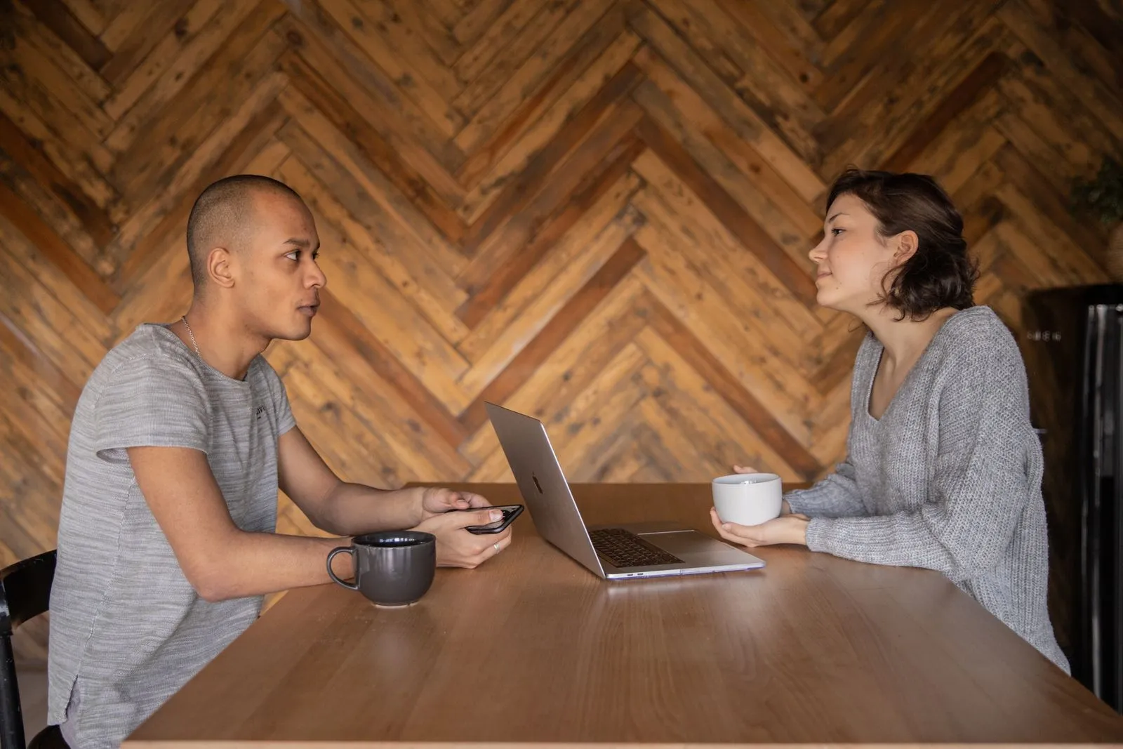 Two people talking across a wooden table with a laptop and mugs