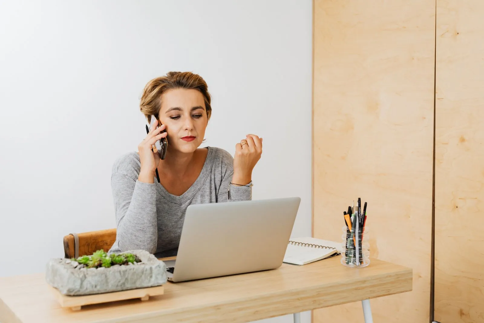 A woman on a phone call at a wooden desk with a laptop