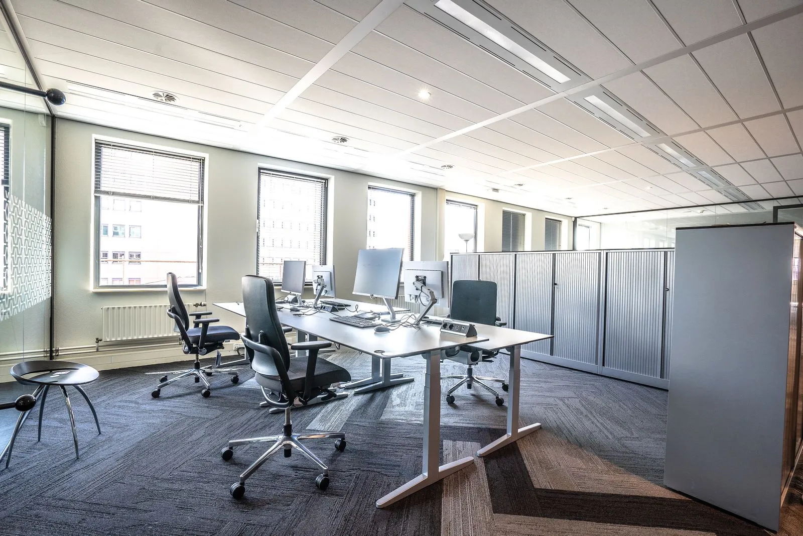 Office workstations with adjustable chairs and monitors beside windows