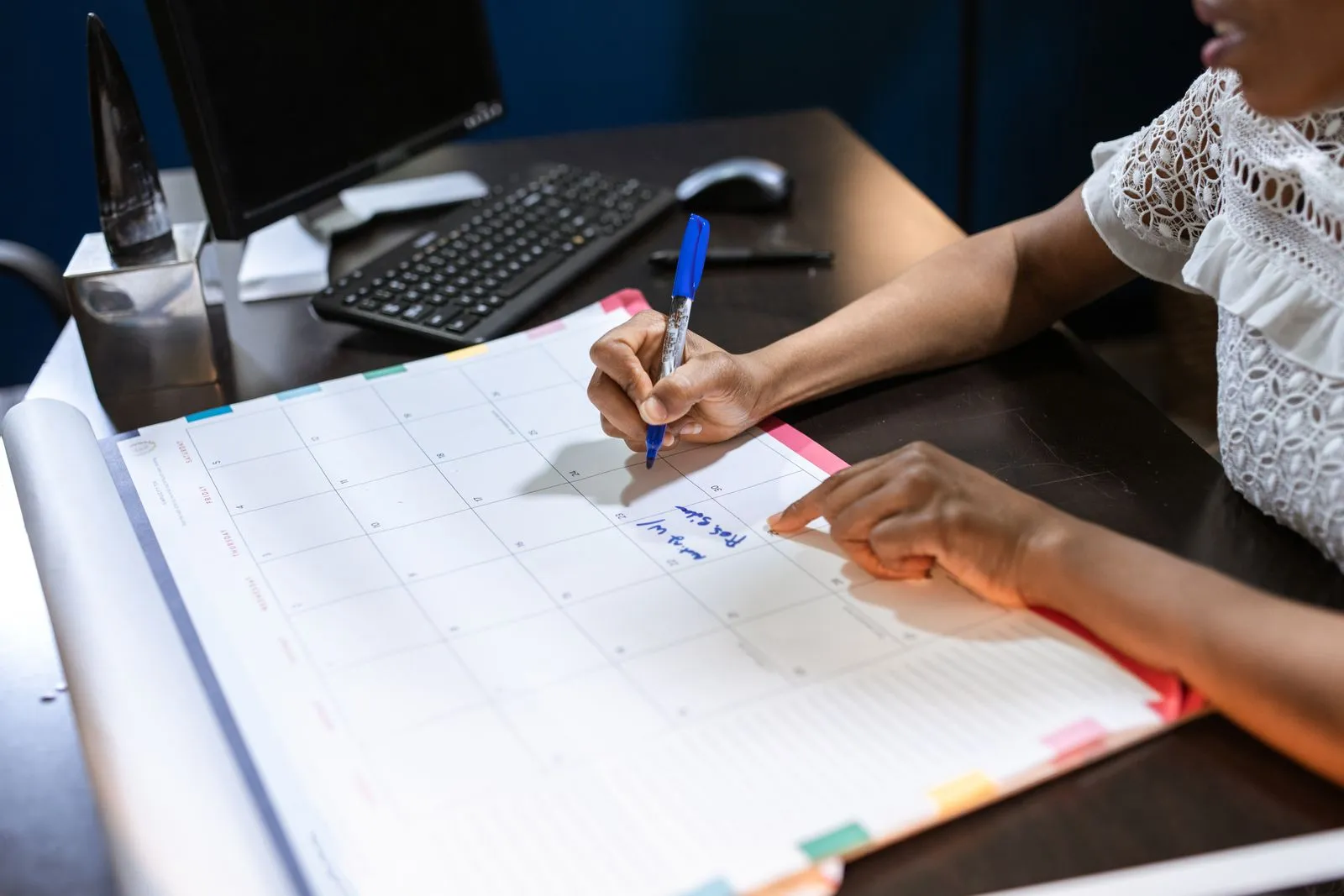 A person writing on a desk calendar beside a computer